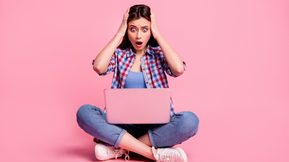 A girl looks in shock at her computer screen on a pink background.