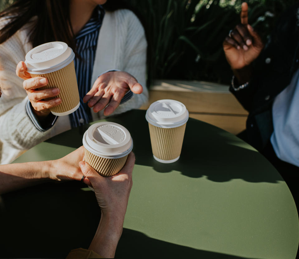 Four people's hands, each holding a paper coffee cup over a table, suggesting a social meeting