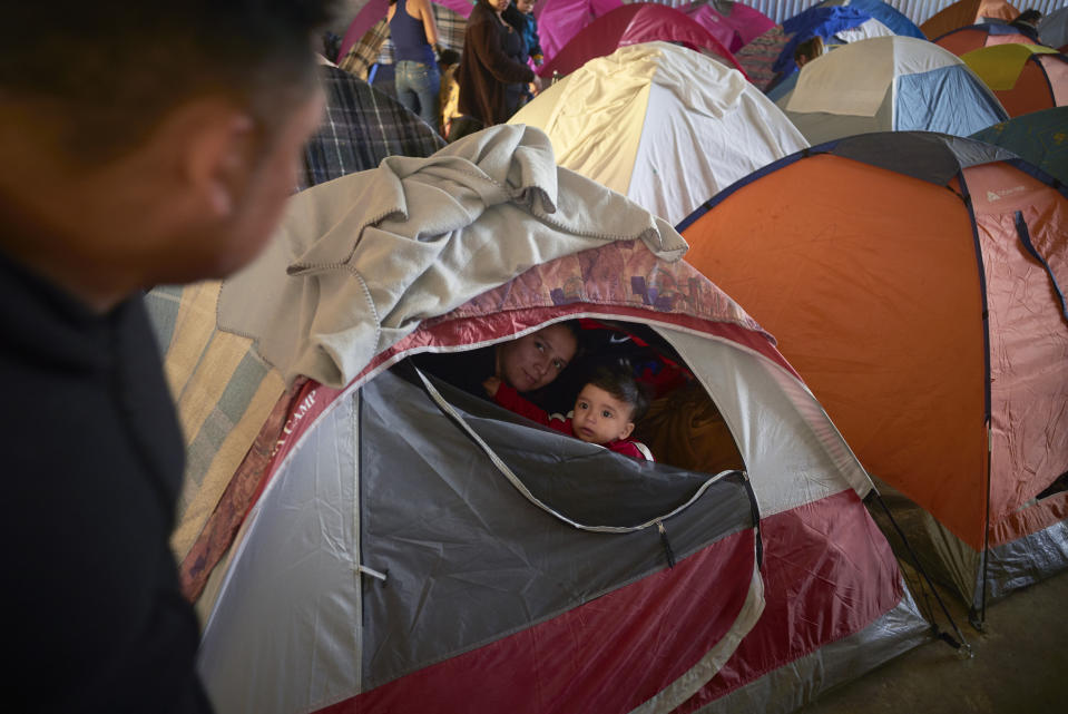 In this March 5, 2019, image, Ruth Aracely Monroy, center, looks out of the family's tent alongside her 10-month-old son, Joshua, as her husband, Juan Carlos Perla, left, passes inside a shelter for migrants in Tijuana, Mexico. After fleeing violence in El Salvador and requesting asylum in the United States, the family was returned to Tijuana to await their hearing in San Diego. They were one of the first families to contend with a new policy that makes asylum seekers stay in Mexico while their cases wind through U.S. immigration courts. (AP Photo/Gregory Bull)