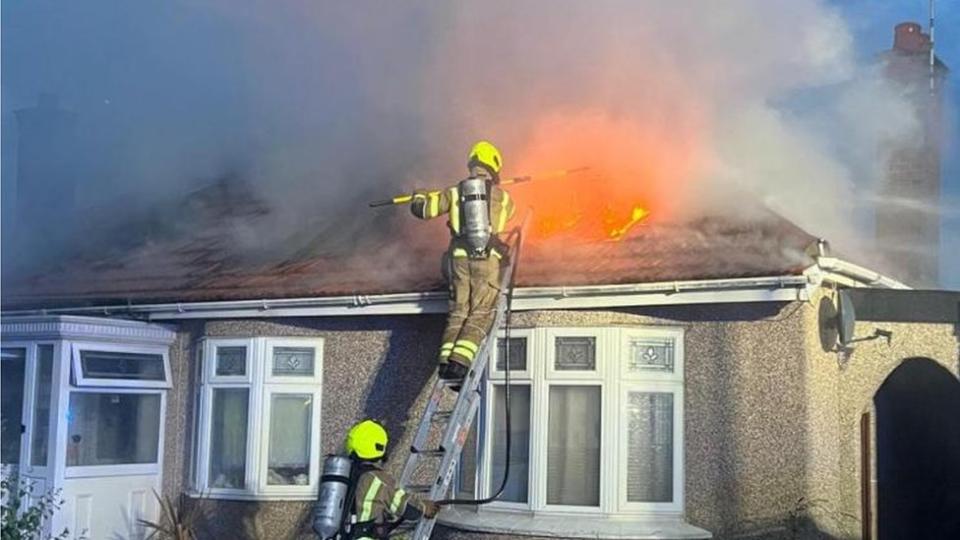Fire fighters up a ladder at the front of a bungalow tacking a fire in the roof