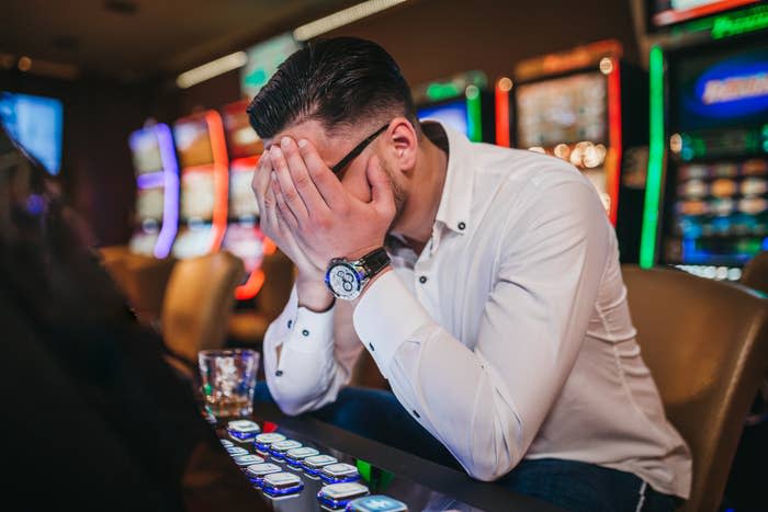 A person wearing a white shirt with their head in their hands, seated in front of a gaming machine in a casino