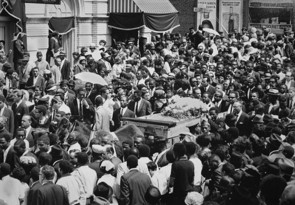 a crowd of people surround a horse drawn cart pulling a casket topped with flowers