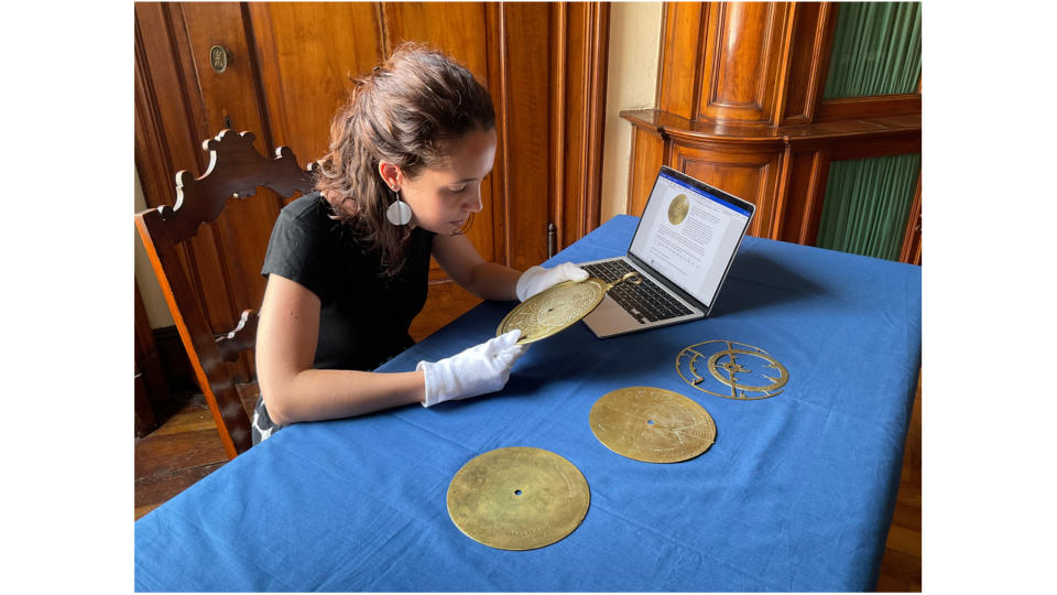 a woman sitting at a table examines a disc-shaped bronze astronomical instrument.