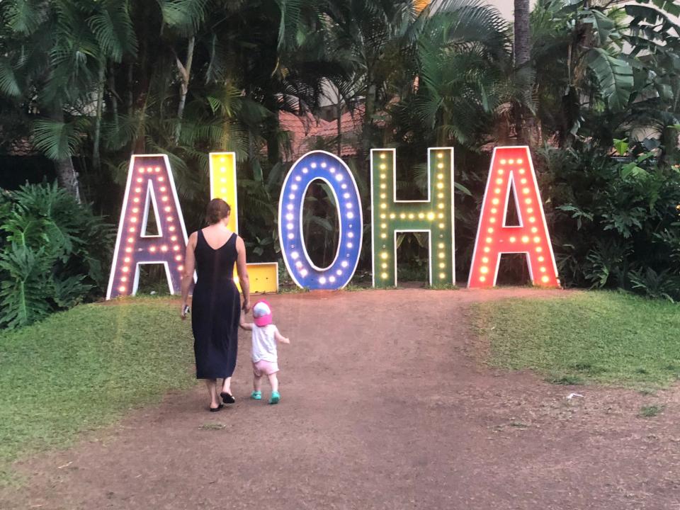 A woman walking towards a light up Aloha sign holding the hand of a child.