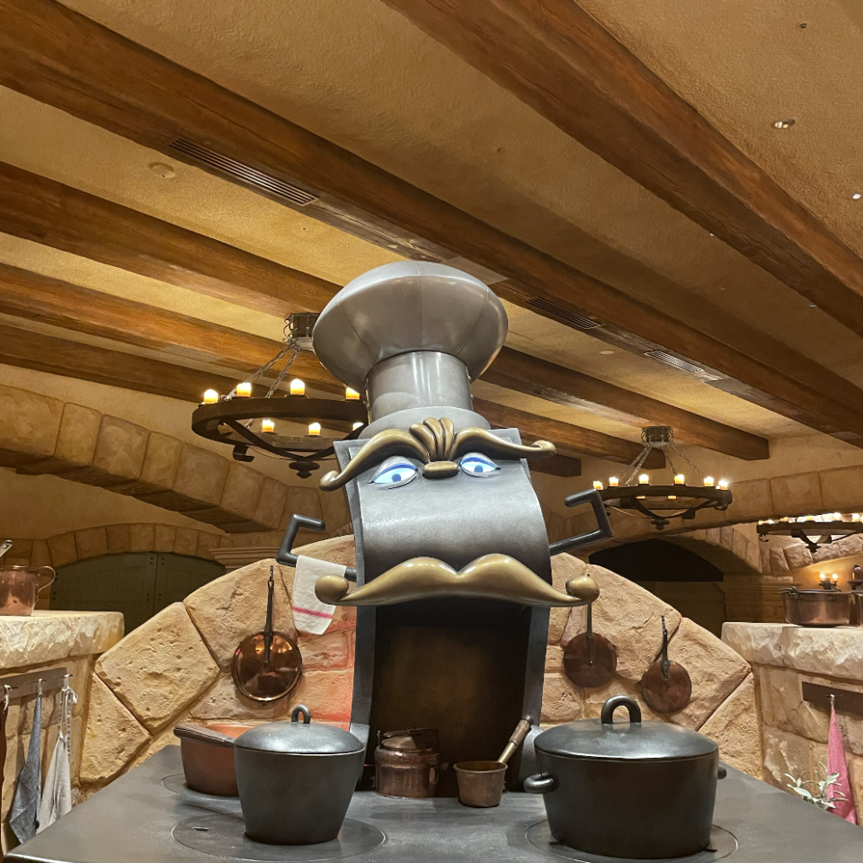 An animatronic chef's stove character from Disney's Beauty and the Beast, set in a rustic kitchen with pots and pans on the stovetop and hanging in the background