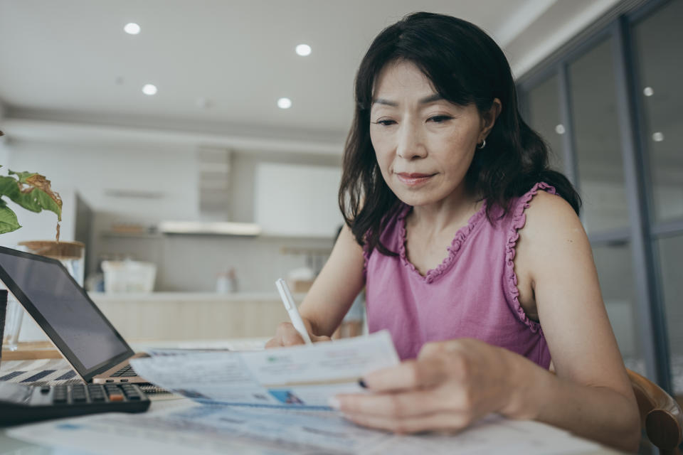 A woman is at home, holding a collection of paper bills and financial plans. She appears to be focused on managing her personal finances, possibly sorting through bills, budgeting, or organizing her financial matters.