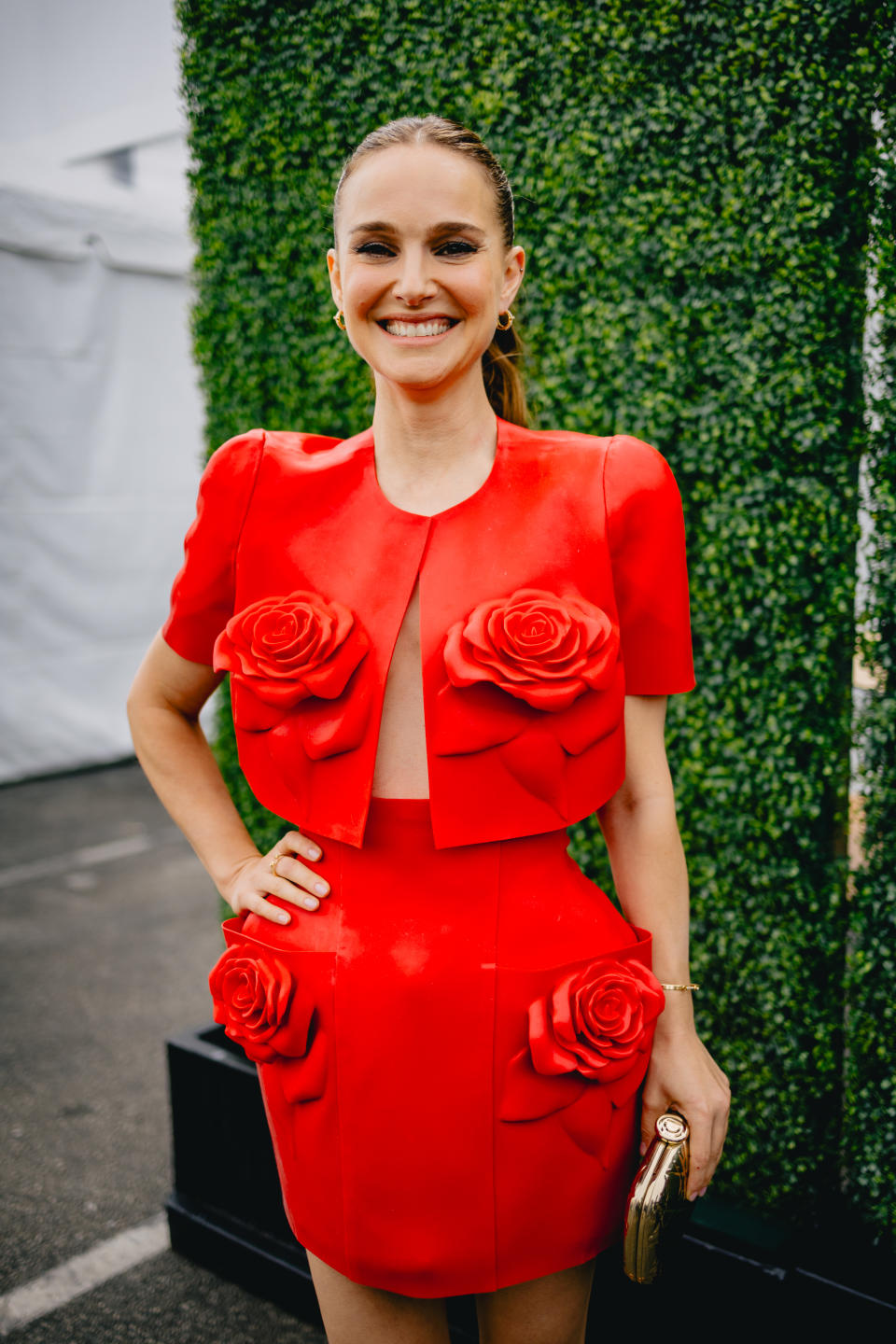 Natalie Portman smiles, wearing a red outfit adorned with large rose designs, posing in front of a green hedge wall