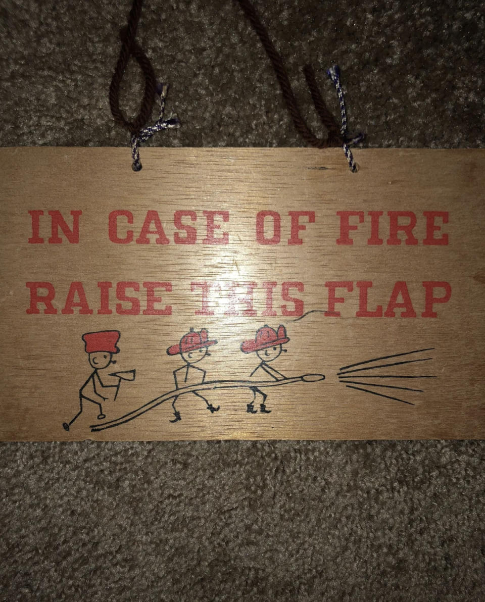 Wooden sign with text "In case of fire raise this flap." Below, three stick figures in fire hats hold a hose spraying water