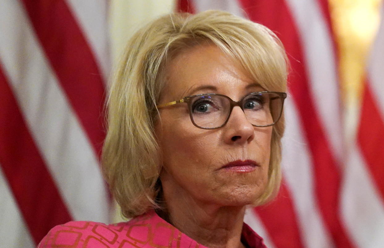 Education Secretary Betsy Devos, with a backdrop of the American flag, looks pensive.