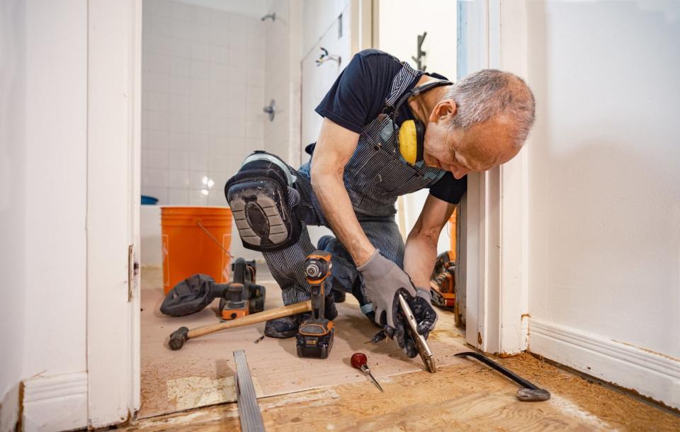 A man in overalls uses tools to fix or replace wood flooring.