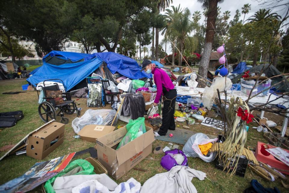 A woman standing near a tent in a park is surrounded by cardboard boxes and plastic bags