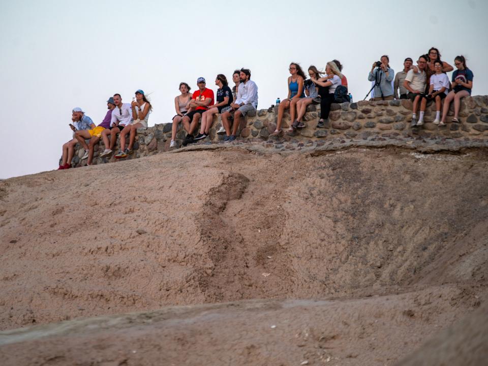 A group of people sitting on a rock on top of a sand dune while looking off in the distance at the rising sun.