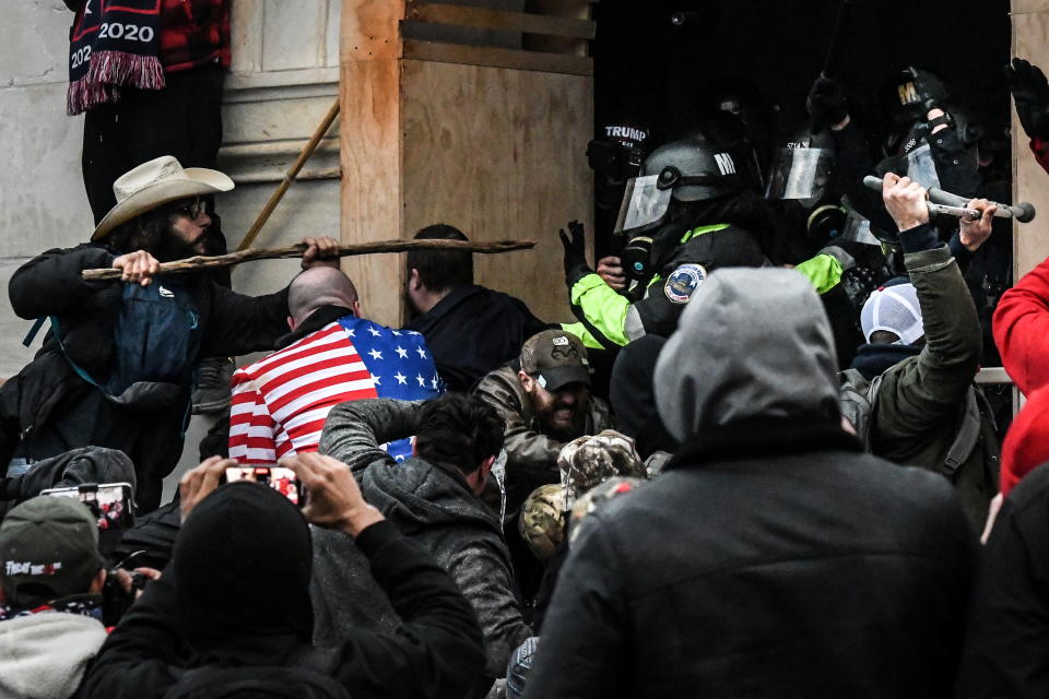 A man in a beige cowboy hat with a tall stick gets ready to batter his way through a door, as riot police in lime-green suits and protective helmets try to prevent a breach as rioters press in around them. Several other people stand by using their cellphones to record the scene.