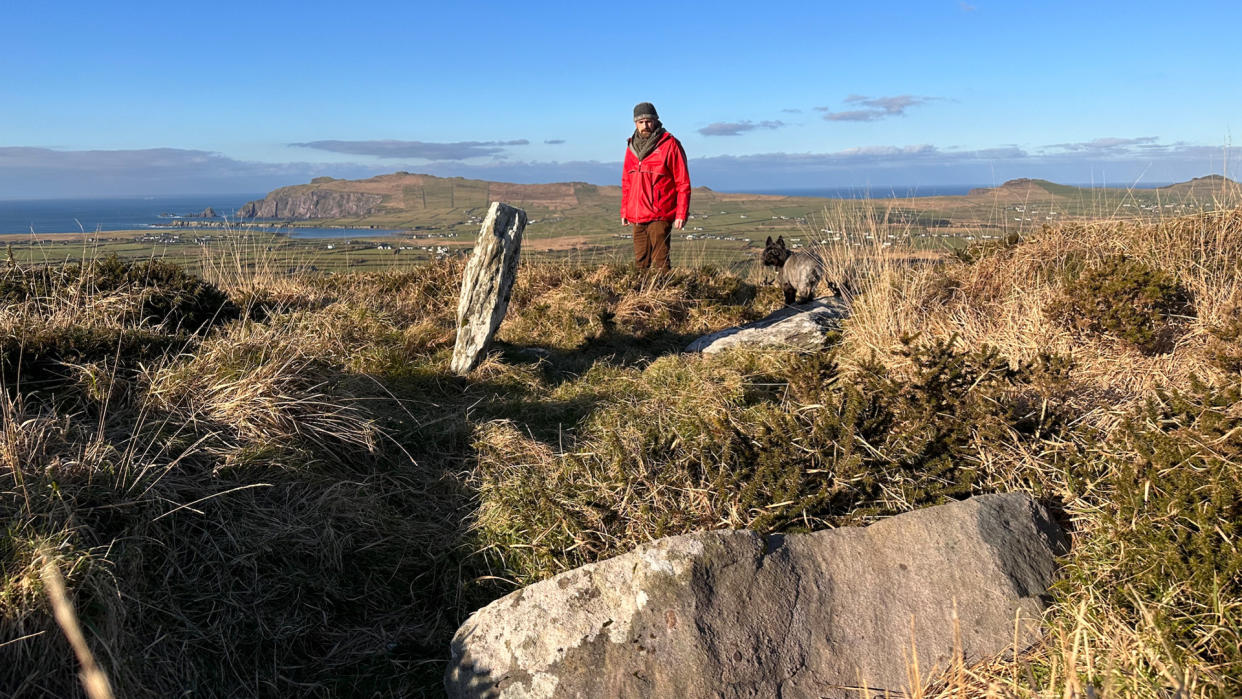  Archaeologist in red coat stands on grassy seaside cliff before a large rock marking a burial site. 