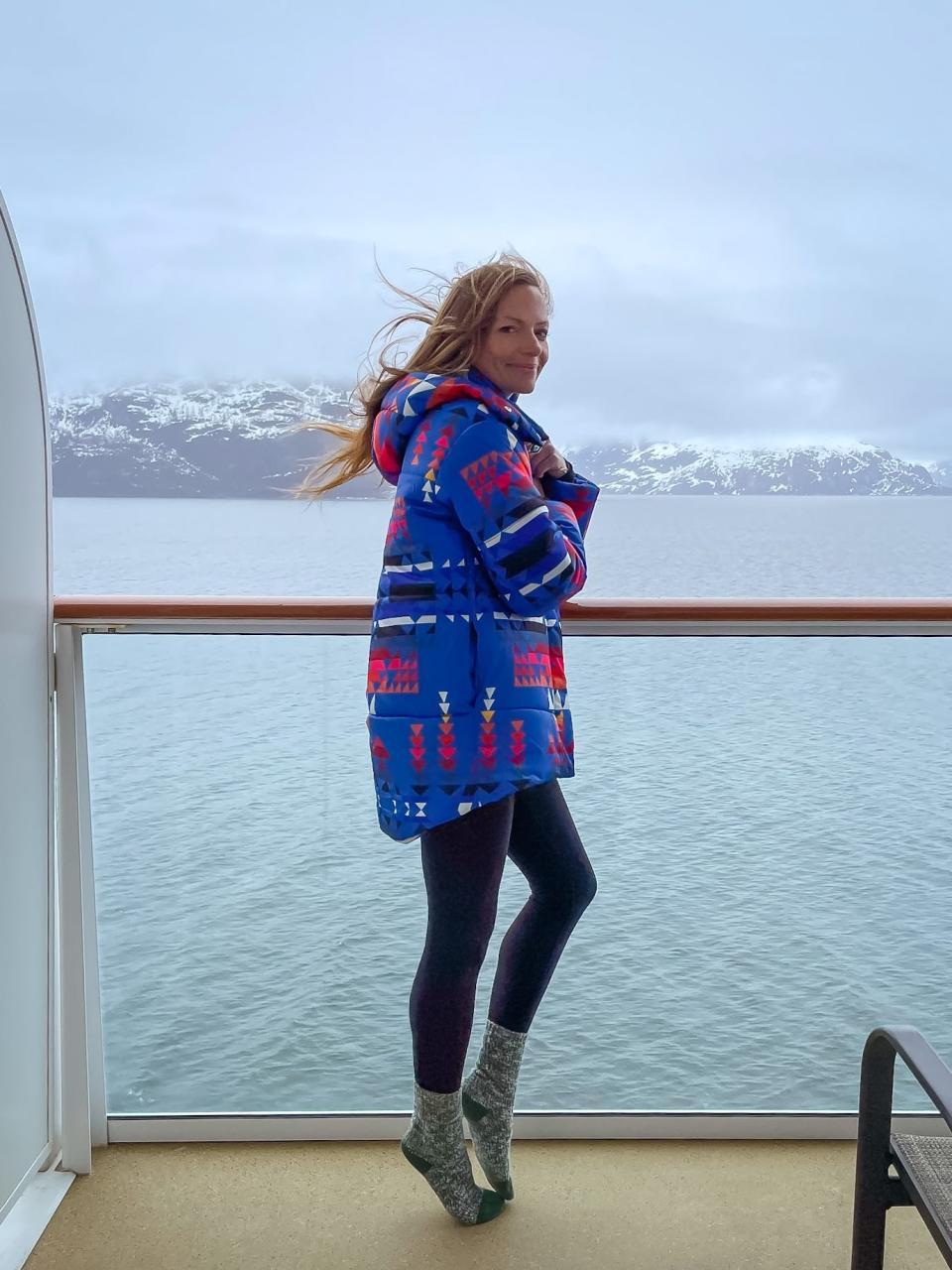 Emily stands on a cruise balcony overlooking glaciers as she wears a heavy coat, black leggings, and grey socks.