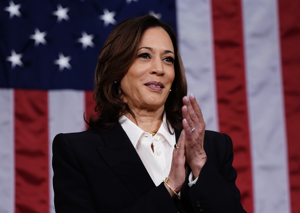 Kamala Harris, wearing a formal suit with a white blouse, claps her hands in front of an American flag backdrop