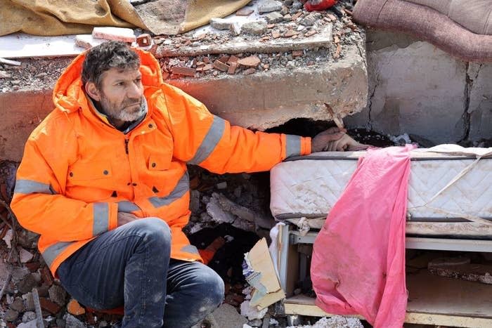 A man in a bright orange jacket sits atop a collapsed building and holds his dead daughter's hand