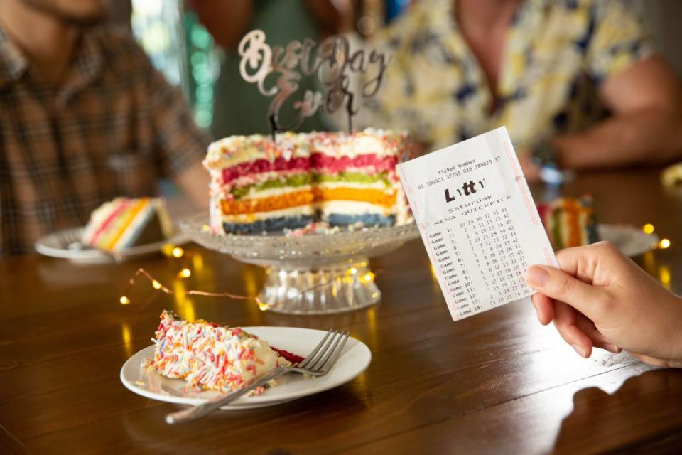 A woman holds a lotto ticket in front of a rainbow birthday cake. 