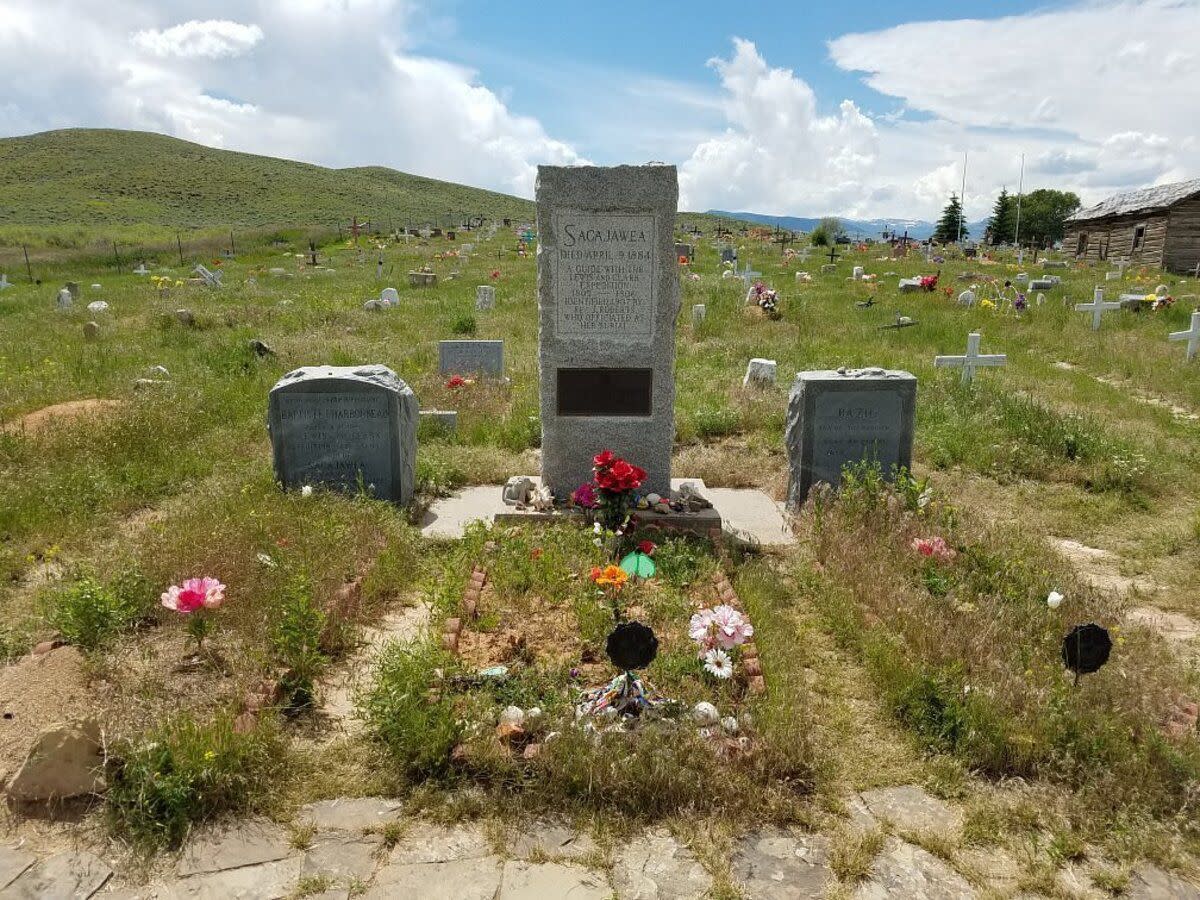 Sacagawea's grave in the the Sacajawea Cemetery, Fort Washakie, Wyoming with flowers in front of it, among several other shoshone tribe graves with hills and a blue sky with white clouds on a summer day