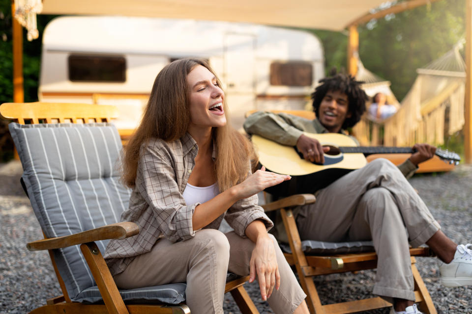 Carefree diverse friends singing and playing guitar, enjoying music near RV on camping trip. Young lady and black guy having fun on summer holidays, spending time in nature