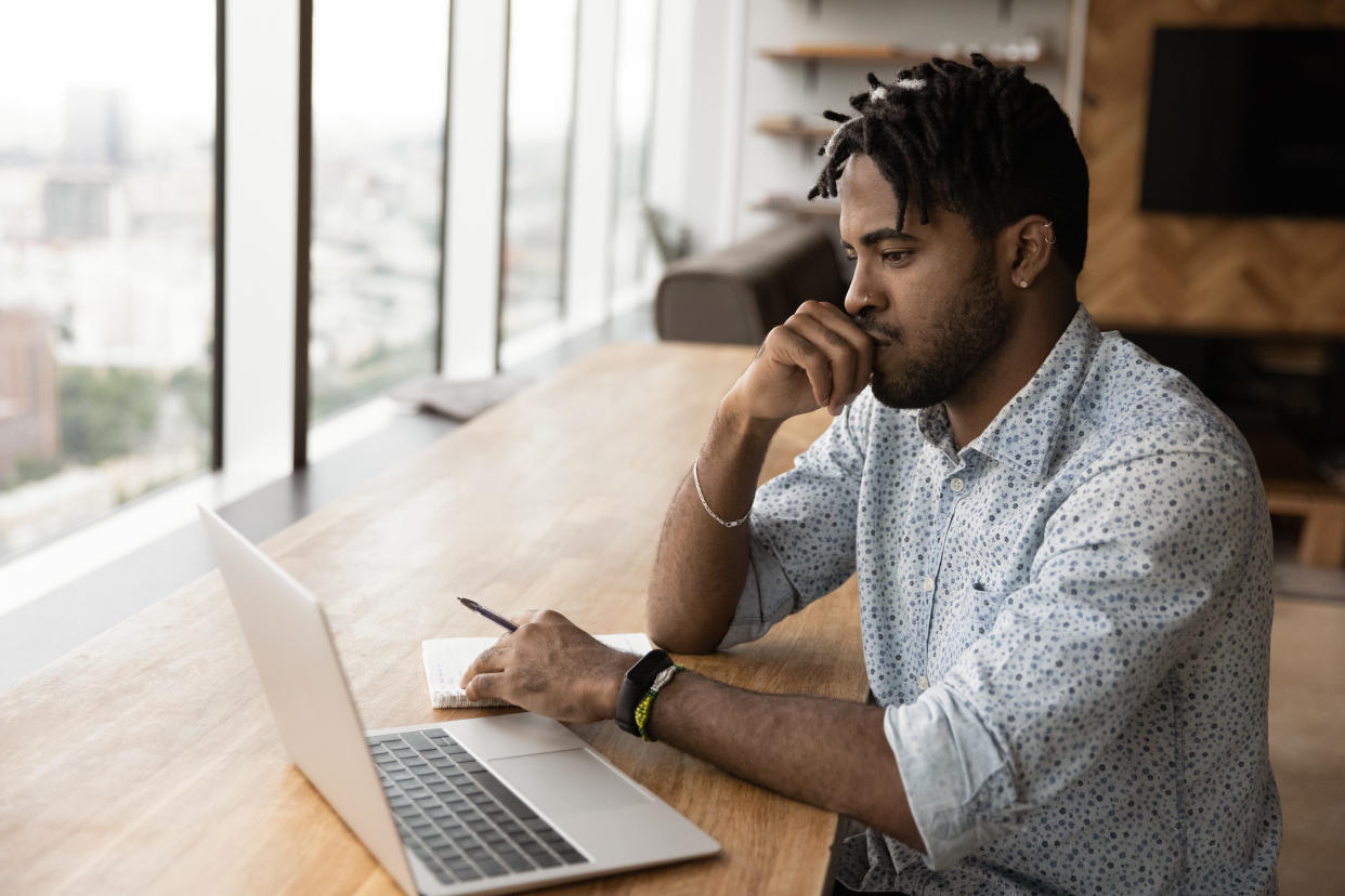 Pensive millennial African guy sit at table looks at laptop screen consider task, e-studying online use modern tech, noting information.