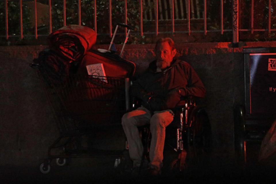 An elderly homeless man sits in his wheelchair next to his belongings on the sidewalk at night.