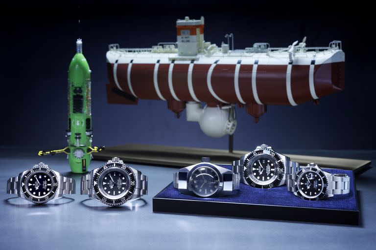 Las diferentes versiones de los relojes  para bajar a la fosa de las Marianas: de izquierda a derecha, los Oyster Perpetual Rolex Deepsea (2012), Oyster Perpetual Deepsea Challenge (2022), Deep
Sea Special (1960), Rolex Deepsea Challenge (2012) y Oyster Perpetual Submariner (1986); atrás de ellos, el sumergible Deepsea Challenge y el batíscafo Trieste