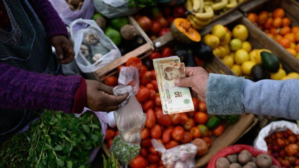 Una persona pagando con pesos argentinos en un mercado de frutas y verduras.