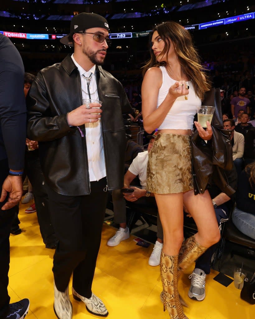 at a basketball game, Bad Bunny in a leather jacket and glasses, Kendall in a snakeskin skirt and knee-high boots