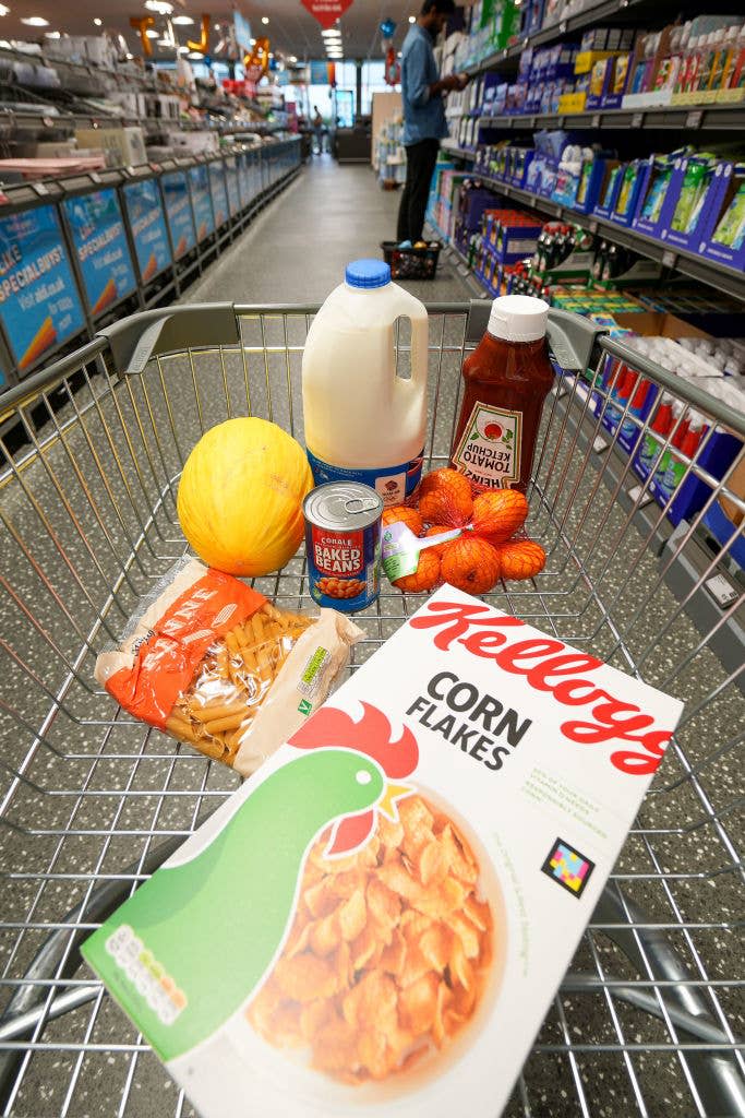 A shopping cart with cereal, pasta, melon, baked beans, milk, ketchup, and strawberries in a supermarket aisle. A person is seen shopping in the background