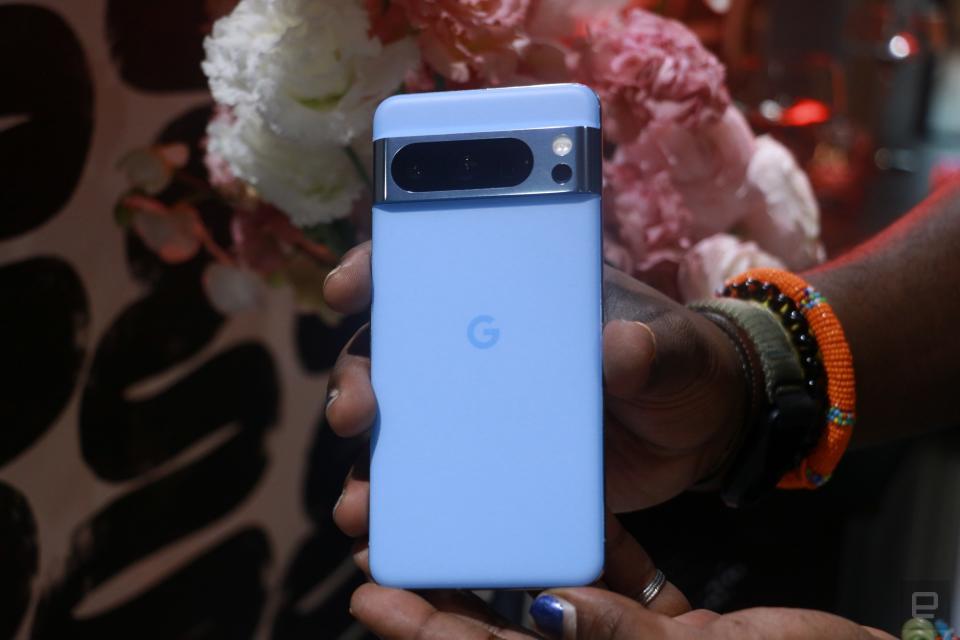 A person holding a blue Pixel 8 Pro in front of some pink and white flowers.