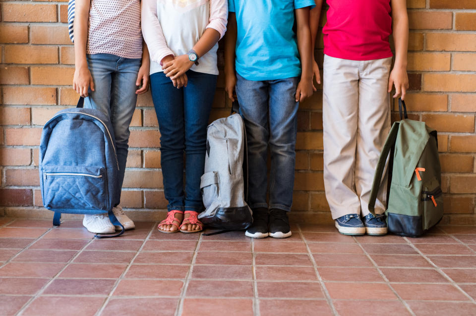 Low section of elementary students standing outside class with backpacks. Legs of four boys and girls leaning in a row. Four multiethnic school children before the start of the lessons.