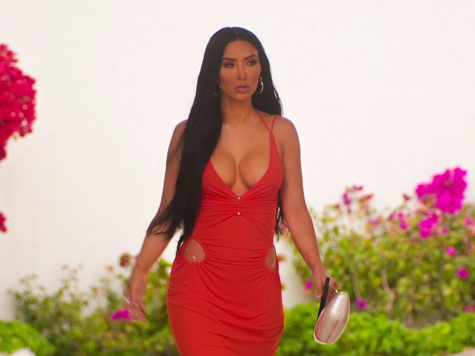 bre tiesi wearing a low cut, form fitting red dress, holding a clutch as she walks through an open door into a home