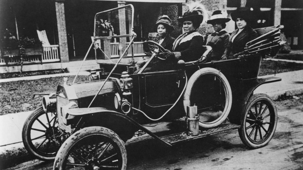 madam cj walker sits in the driver seat of an early car with the top down, a woman sits in the passenger seat, both women wear hats