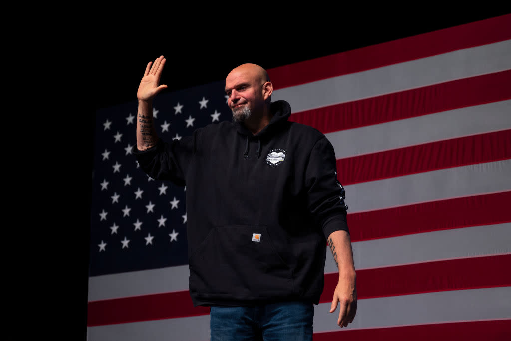 John Fetterman waves his hand as he stands behind an American flag.