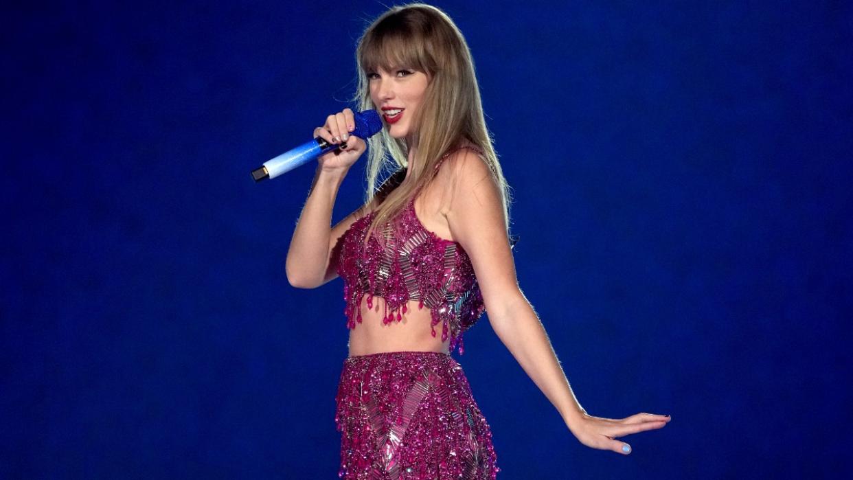  Taylor Swift holding up a mic and putting her hand out while singing in a pink two-piece set on the Eras Tour. 