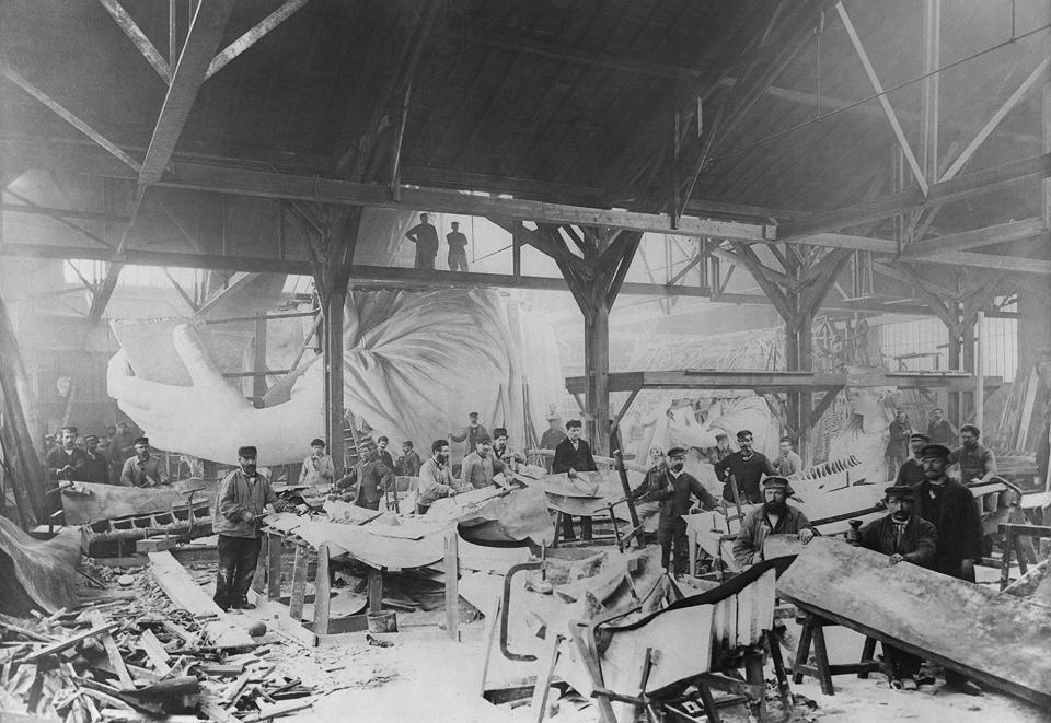 Men standing around the interior of an edifice with lots of debris around and the statue's arm visible on one side