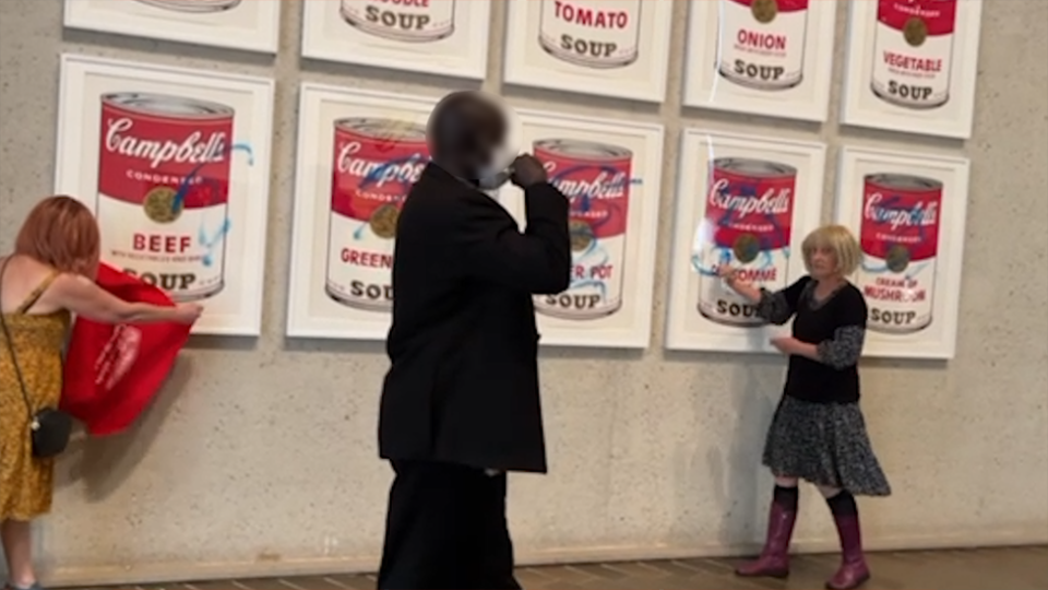 Activists scrawling blue letters on Campbell's soup can. A security guard is in the foreground.