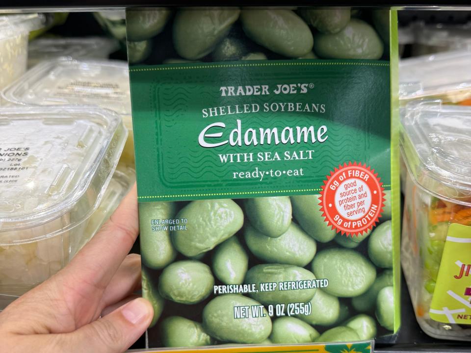 Hand holding Trader Joe's edamame in front of the refrigerator section