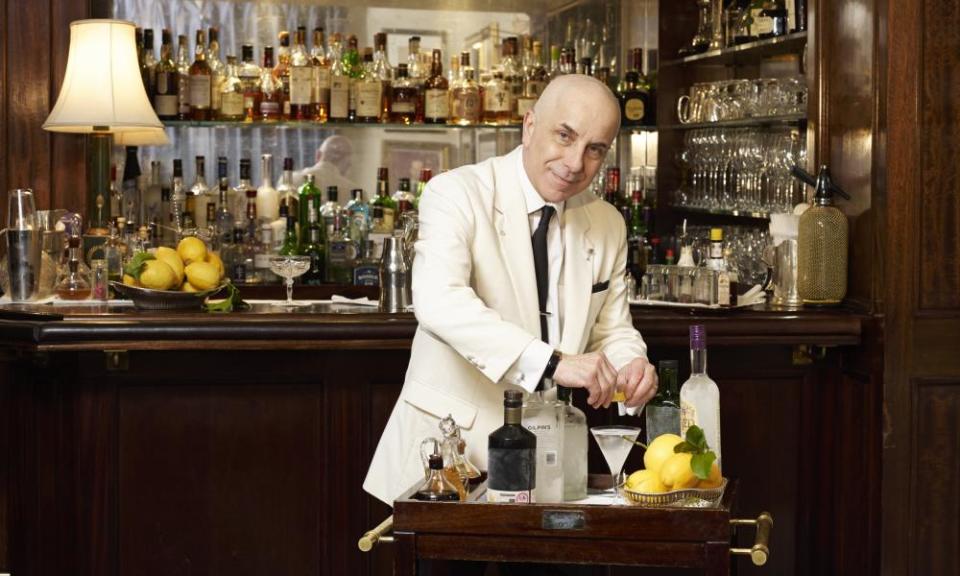 Smiling man in white jacket and black tie prepares a martini at a trolley in front of a bar