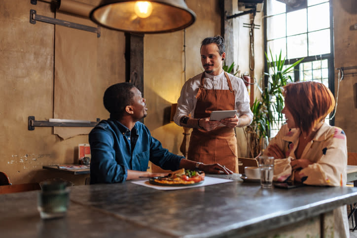 Waiter in an apron serving food to a seated man and woman at a restaurant table