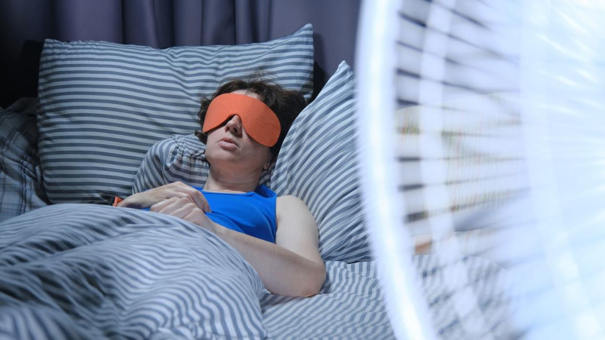 woman is using eye mask in bed while electric fan is blowing on the foreground
