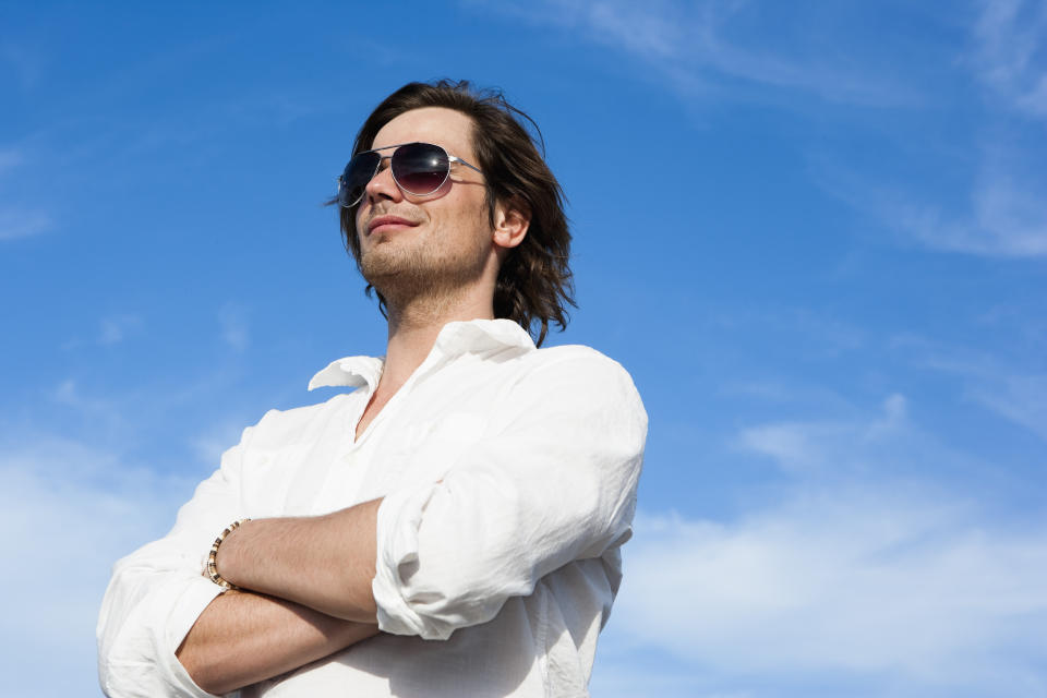 A person wearing sunglasses and a white shirt stands confidently with arms crossed, looking away against a blue sky