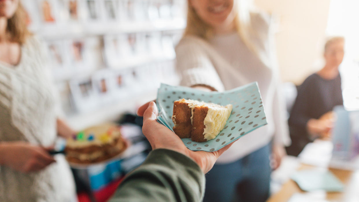 A young woman at her office desk with a birthday cake and handing a slice to a work colleague.