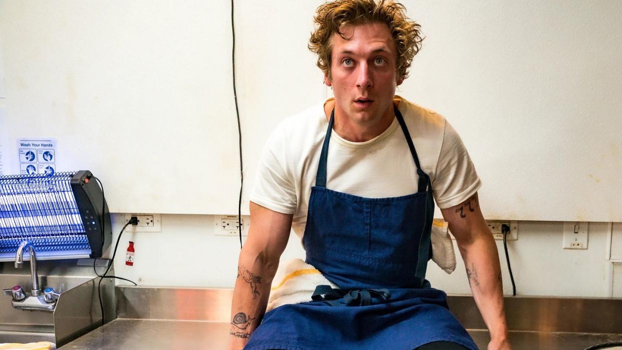 the bear, jeremy allen white sitting on top a metal work surface in a blue chef apron, looking stressed