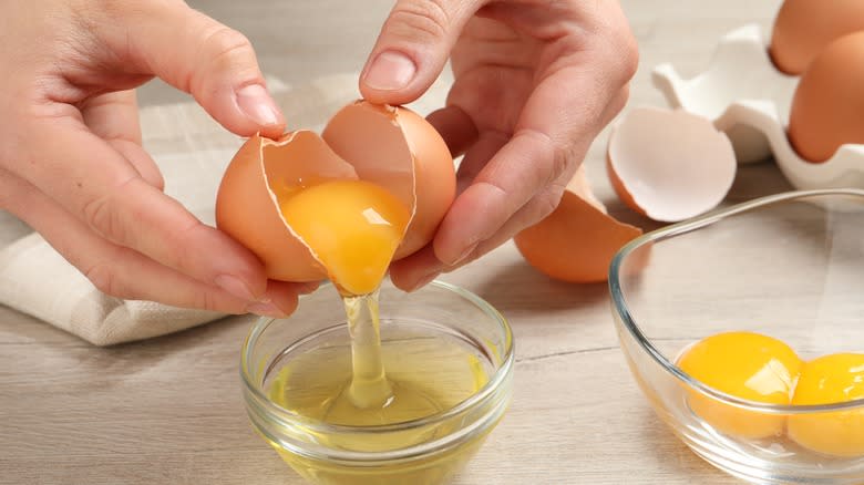Hands separating an egg yolk from a cracked egg over a glass bowl