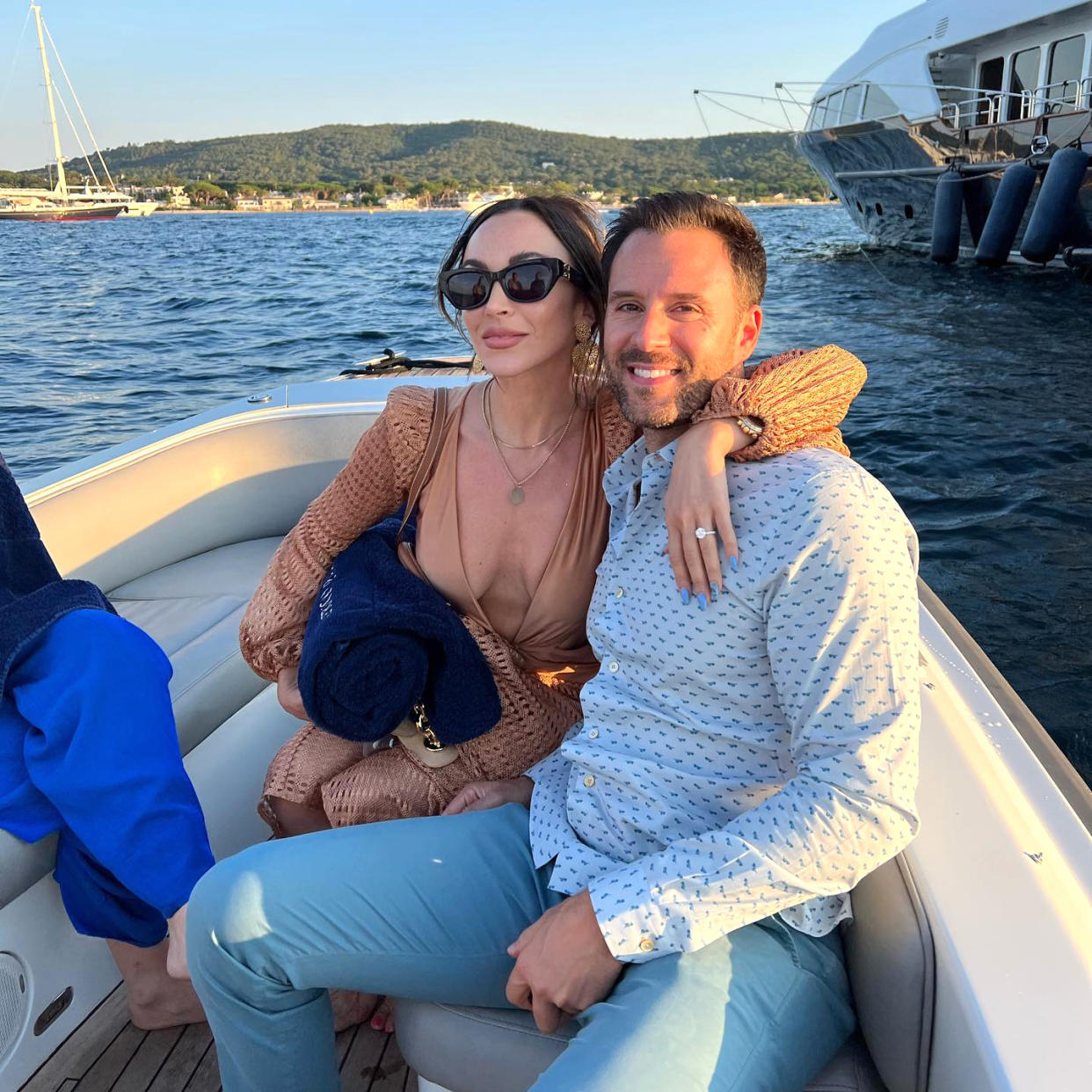 Farrah Aldjufrie in deep V neck beige dress and black sunglasses puts her arm around Alex Manos wearing a button down while riding on a boat with water in background