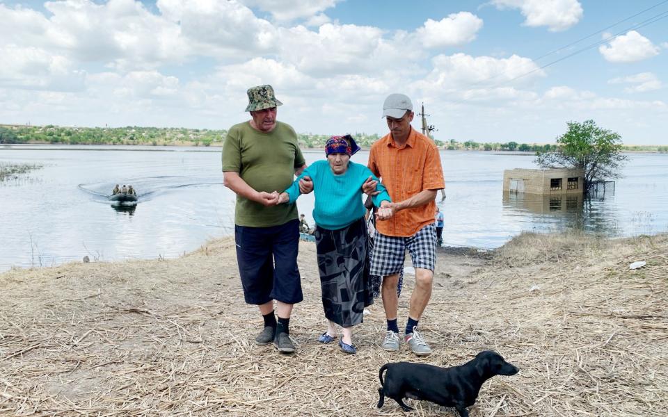 Two men assist an elderly woman as she is evacuated from a residential area flooded by the destruction of the Kakhovka dam. A little dog walks in front of them as they help her to walk away from the flood waters behind them