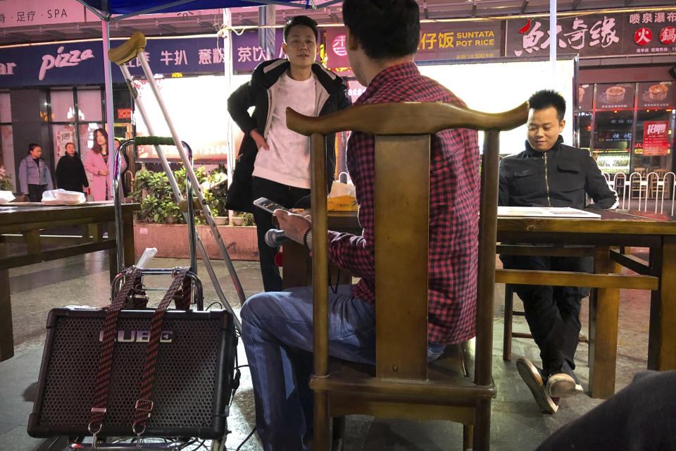 In this Dec. 6, 2019, photo, passers-by talk to Wu Yi, who has struggled with Oxycontin abuse, as he waits for a break in the rain while making his rounds to sing songs for money at all night-restaurants and clubs in Shenzhen in southern China's Guangdong Province. Officially, pain pill abuse is an American problem, not a Chinese one. But people in China have fallen into opioid abuse the same way many Americans did, through a doctor's prescription. And despite China's strict regulations, online trafficking networks, which facilitated the spread of opioids in the U.S., also exist in China. (AP Photo/Mark Schiefelbein)