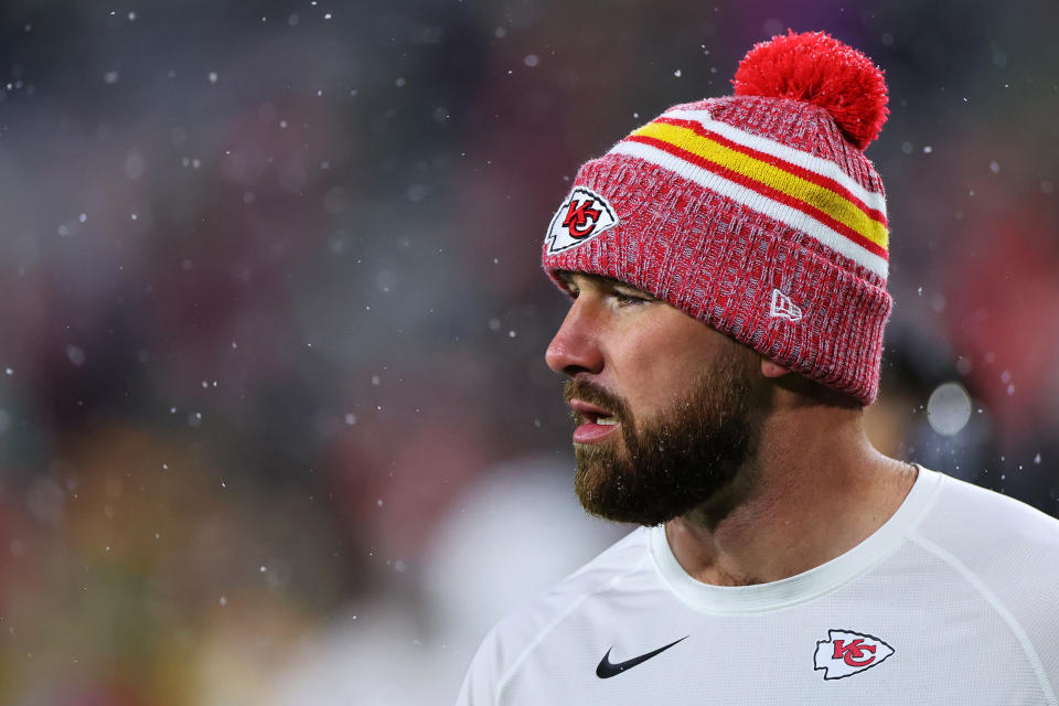 A man with a beard, wearing a Kansas City Chiefs beanie with a pom-pom and a white Chiefs shirt, is seen in a snowy outdoor setting